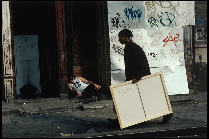 This is a still from the film 'Downtown 1981,' featuring Jean-Michel Basquiat carrying a painting.